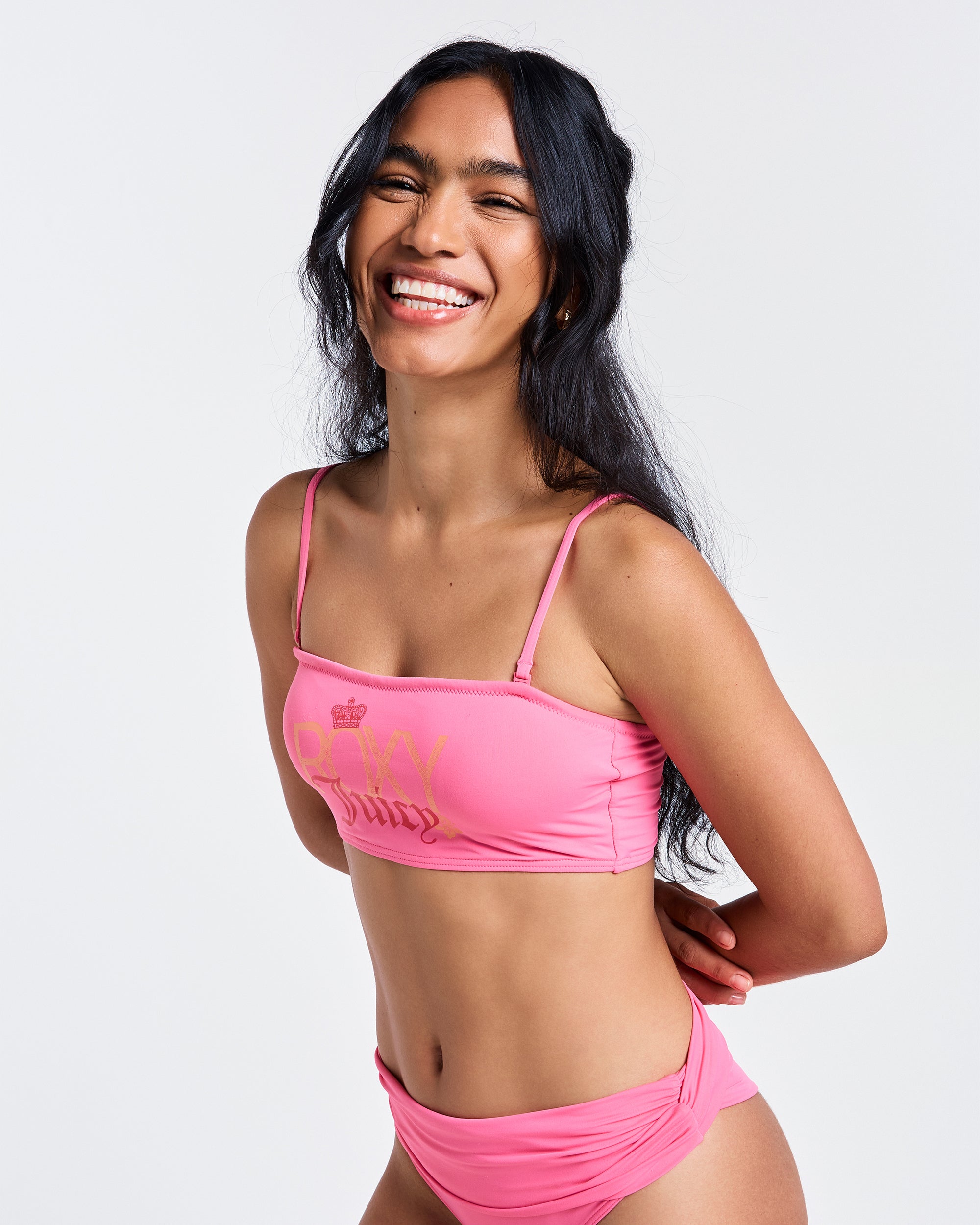 A woman with long dark hair smiles brightly in the Roxy x Juicy Bandeau Bikini Top by Roxy, made from recycled nylon in Azalea Pink, standing against a plain white background.