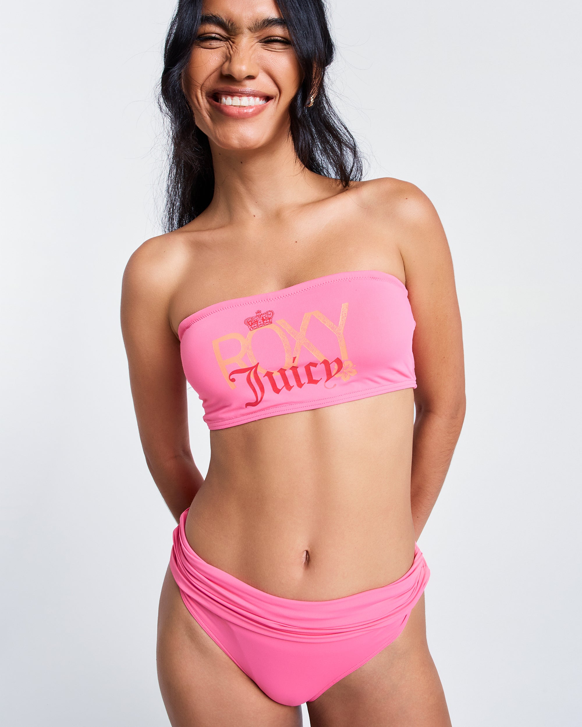 A person smiles in a bright Azalea Pink Roxy x Juicy Bandeau Bikini Top by Roxy, made from recycled nylon, with matching bottoms, standing against a plain light background.