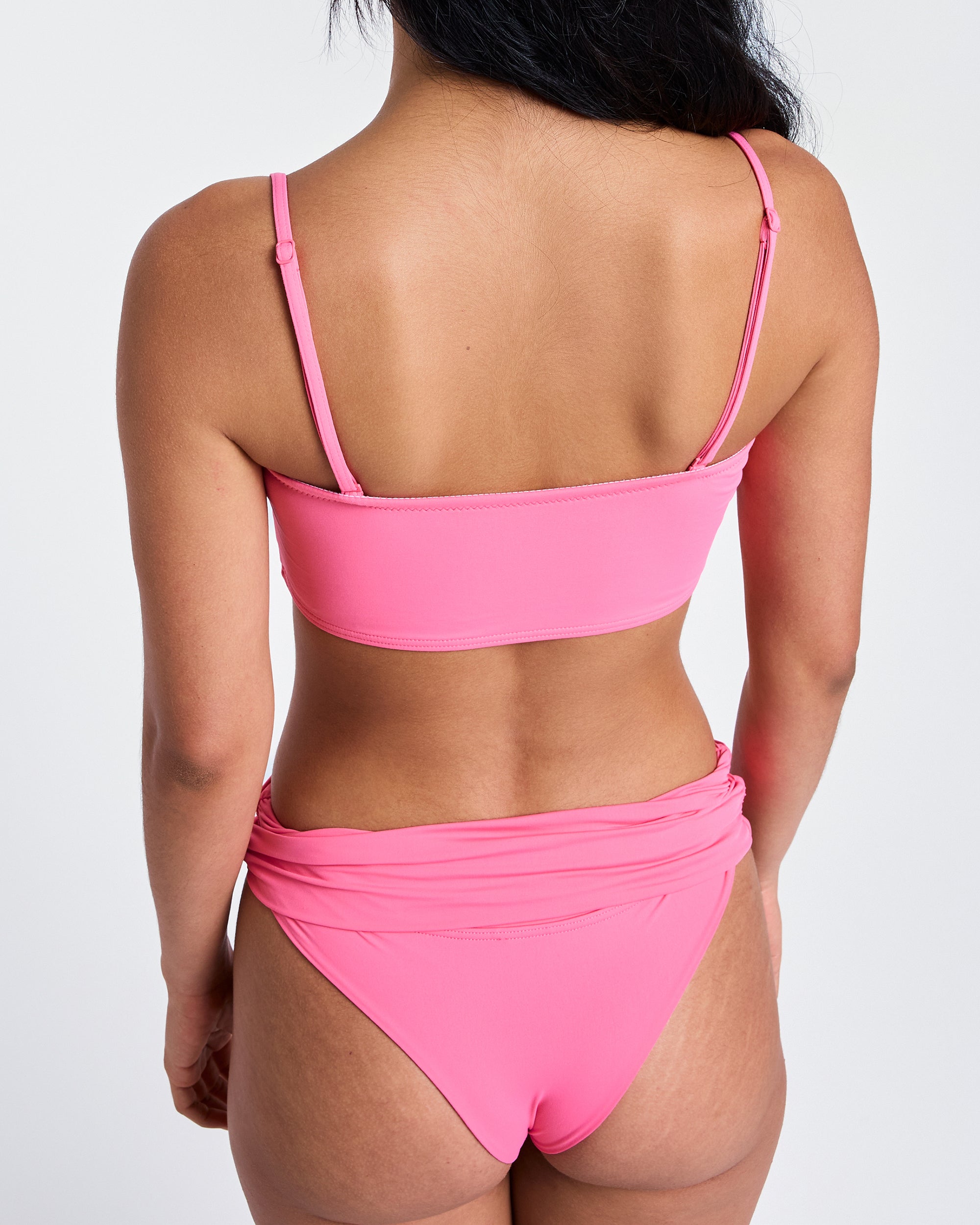 A woman faces away from the camera in an Azalea Pink bandeau top and Roxy x Juicy Cheeky Bikini Bottom by Roxy. Her long dark hair falls over one shoulder, highlighting the surf-inspired style against a plain white background.