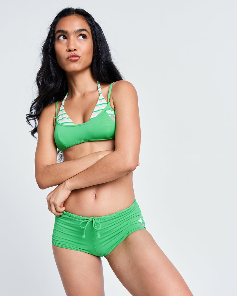 A woman with long dark hair wears the Roxy x Juicy Cheeky Shorty Bikini Bottom, arms crossed as she gazes thoughtfully upward against a plain background.