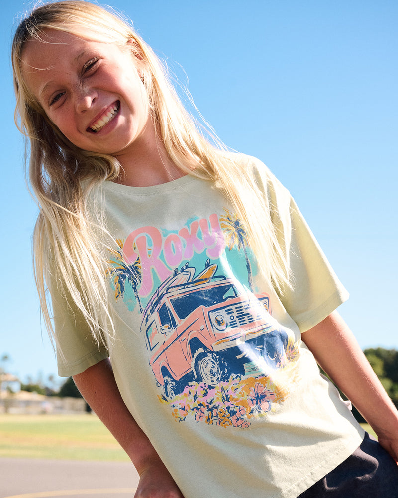 A smiling child with long blonde hair wears the Roxy Girls' Coastal Oversized Tee in Laurel Green, featuring a colorful graphic of a vintage car, surfboards, and palm trees, standing outdoors on a sunny day.