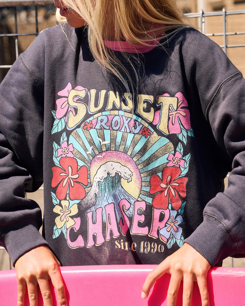 A person holding a pink surfboard wears the Roxy Girls' Sunny Crew, an oversized cotton-poly fleece with a colorful “Sunset Chaser Roxx since 1990” graphic featuring a wave, sun rays, and red flowers.