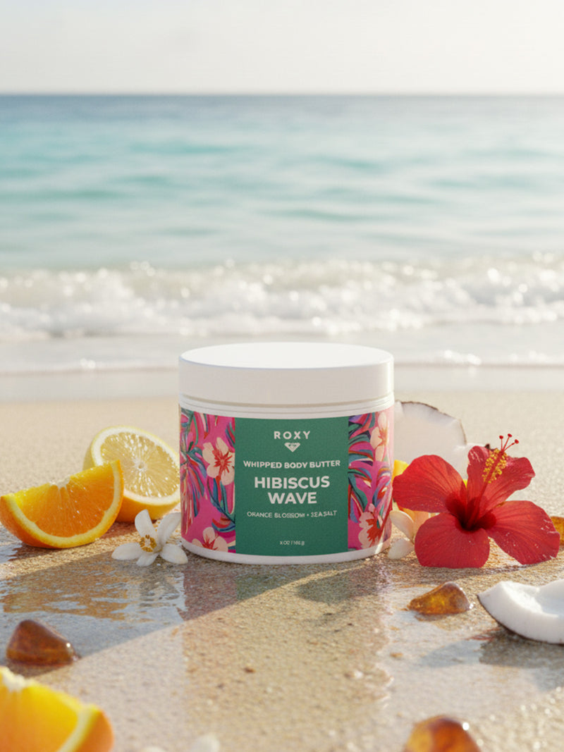 A jar of Roxy Hibiscus Wave Body Butter rests on sandy beach with waves behind, surrounded by orange slices, coconut, and hibiscus—ideal for after-shower moisture and soothing dry skin.