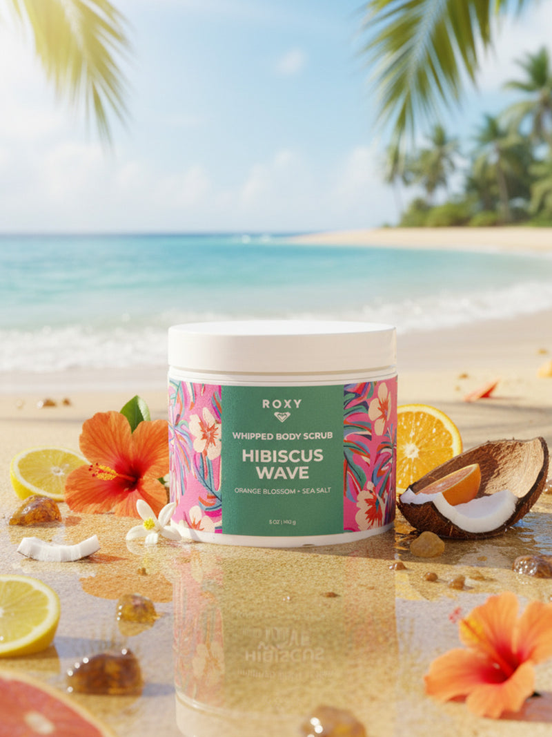 A jar of Roxy Hibiscus Wave Body Scrub rests on a sandy beach with hibiscus flowers, orange slices, and coconut pieces—ideal for exfoliating and giving your shower routine a fresh tropical glow.