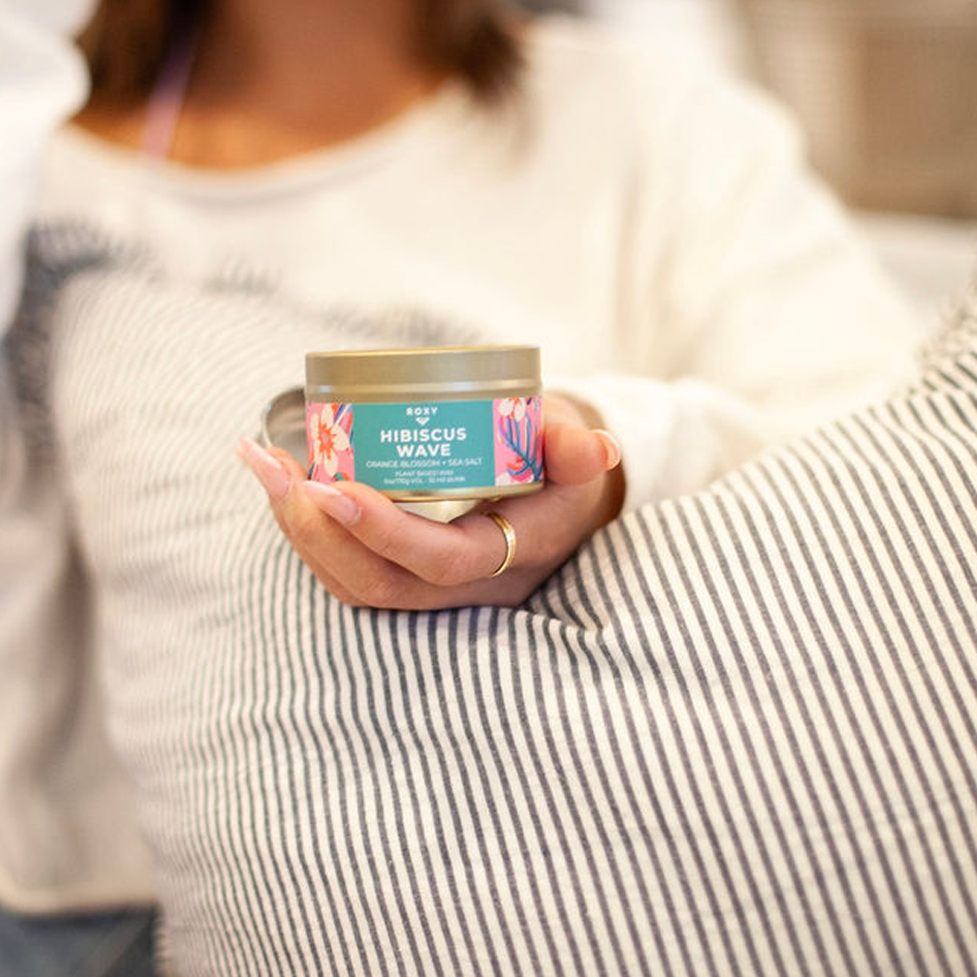 A person in a white shirt holds the Roxy Hibiscus Wave Palm Tin Candle with a vibrant floral design, suggesting tropical scents, while their hand rests on a striped pillow. Their face is out of focus.