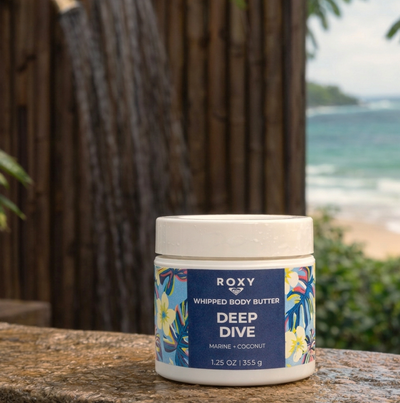 A container of Roxy Deep Dive whipped body butter sits on a stone surface outdoors, with tropical plants and a glimpse of the ocean and sandy beach in the blurred background.