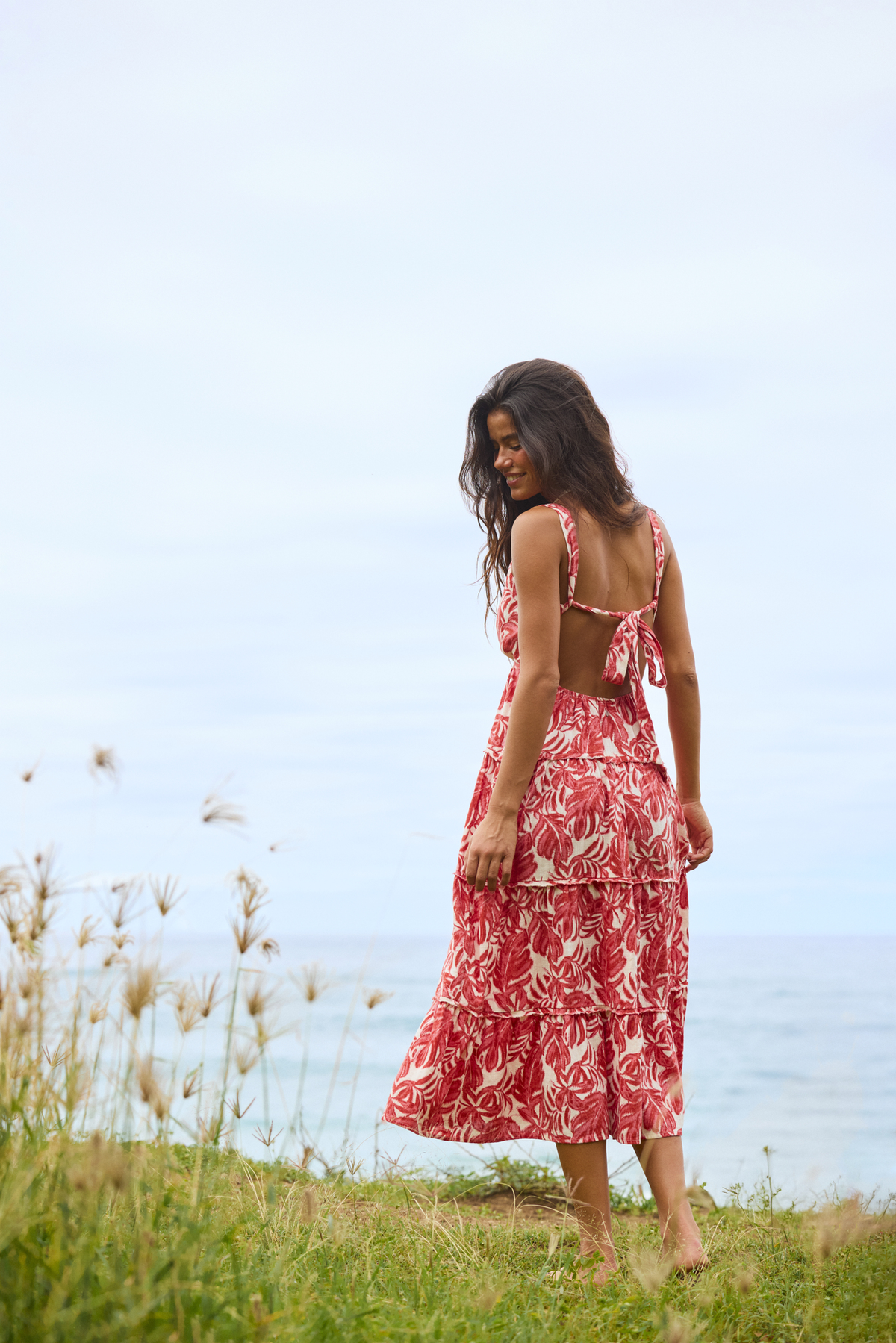 A woman wearing a red and white tropical floral print maxi dress stands on a grassy hill overlooking the ocean, seen from behind.