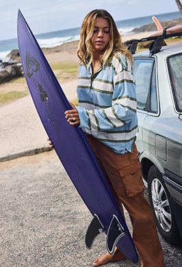 A woman standing barefoot on a beach next to a car, holding a purple surfboard. She is wearing a light blue and gray striped long-sleeve shirt and brown cargo pants.