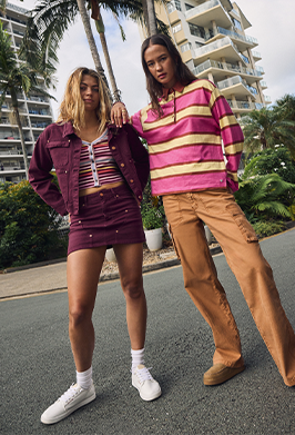 Two models posing outdoors in a tropical city setting, showcasing coordinated streetwear outfits. One model wears a purple denim jacket and mini skirt with a striped crop top, while the other wears a pink and yellow striped long-sleeve top with tan cargo pants.