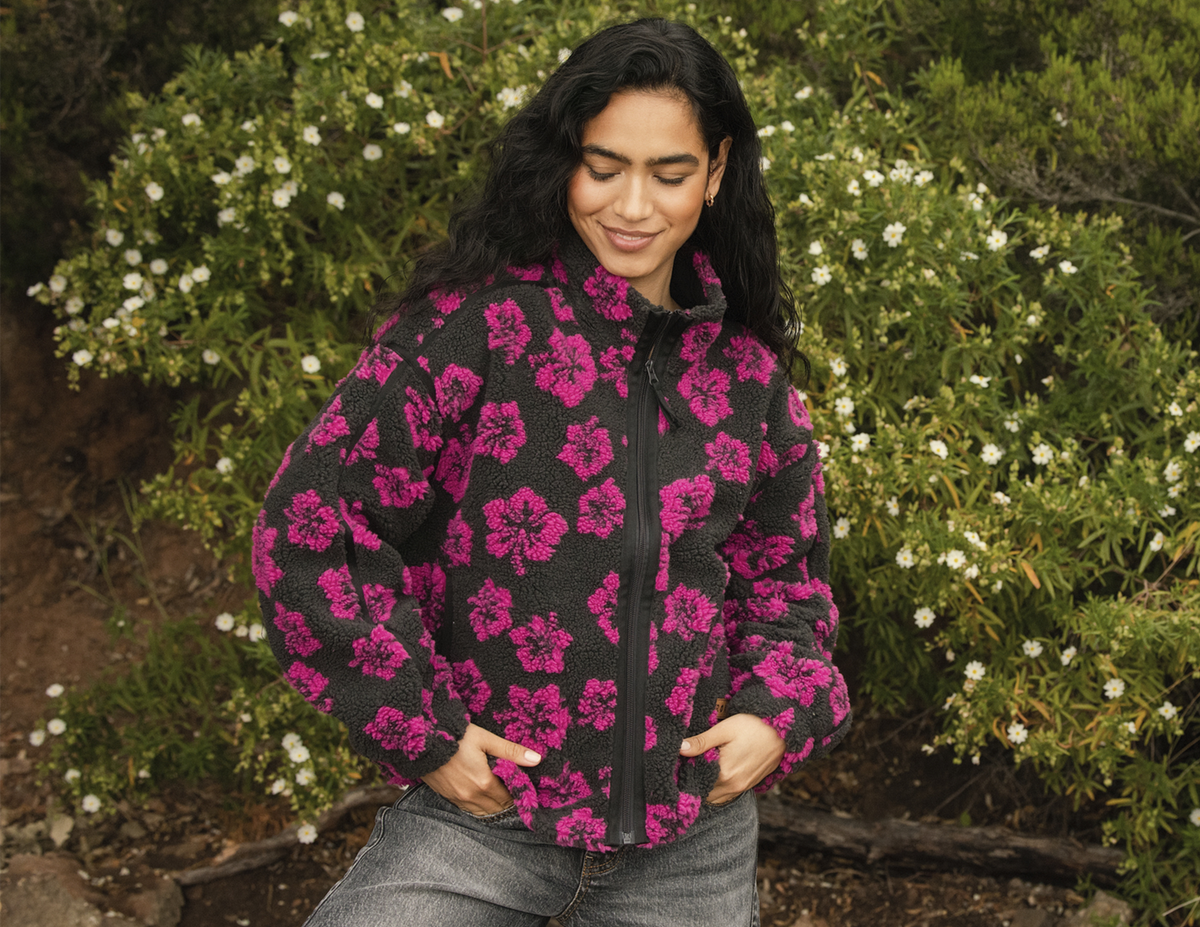 A woman wearing a black fleece jacket with a vibrant pink floral pattern, standing outdoors in front of green bushes with small white flowers.