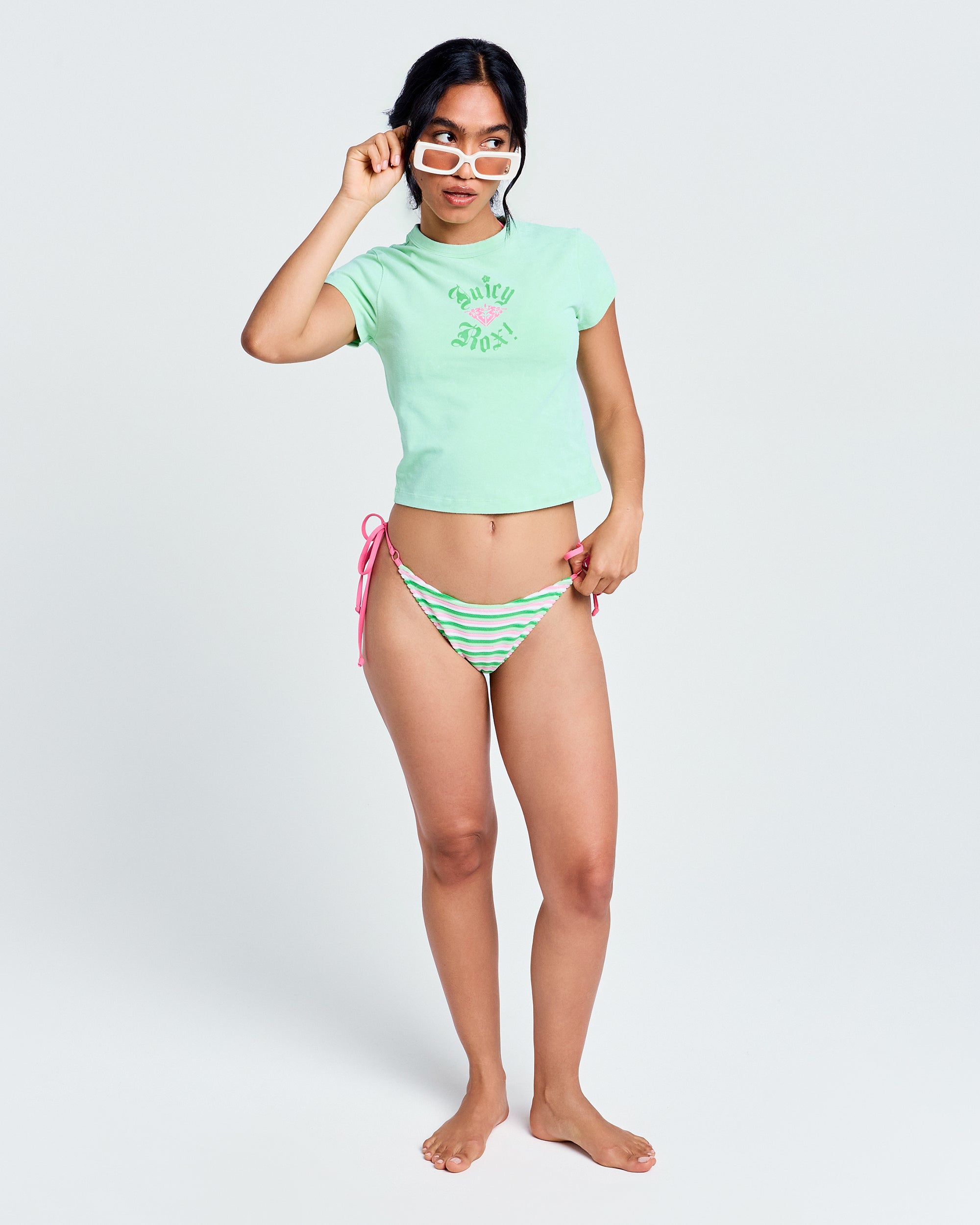 A woman poses on a light background wearing a fitted short sleeve Roxy x Juicy Baby Tee in Green Ash, striped bikini bottoms with pink side ties, and white-framed glasses, holding the glasses with one hand and pointing with the other.