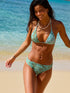 A woman in the Roxy Sea Bloom Mini Tiki Triangle Bikini Top with adjustable tie straps smiles on a sandy beach by clear blue water. Her hair is wet, and she enjoys a sunny seaside day.