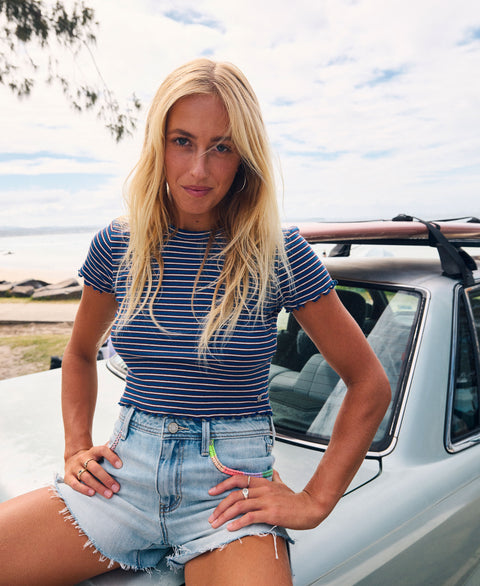 A woman with long blonde hair, dressed in the Roxy Poolside Crop Tee and denim shorts, sits on the hood of a light blue car by the beach, looking at the camera on a bright, sunny day and effortlessly showcasing summery flair.