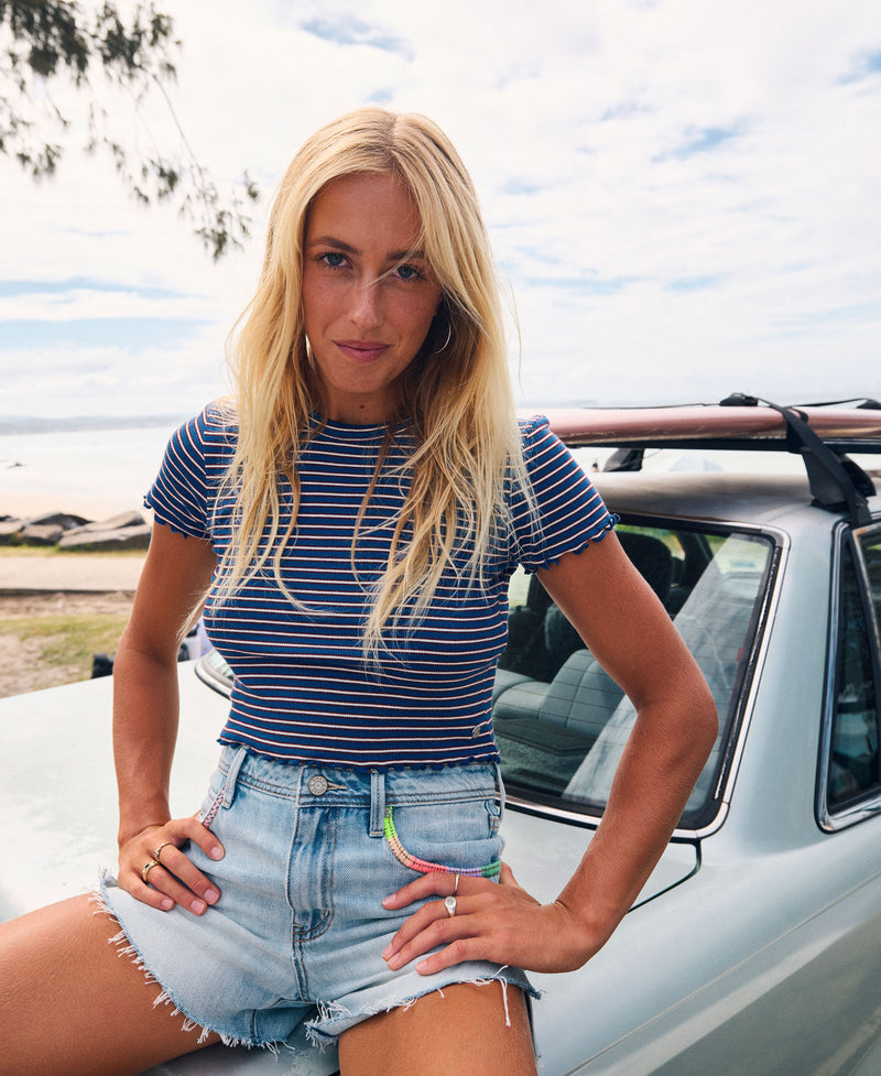 A woman with long blonde hair, dressed in the Roxy Poolside Crop Tee and denim shorts, sits on the hood of a light blue car by the beach, looking at the camera on a bright, sunny day and effortlessly showcasing summery flair.