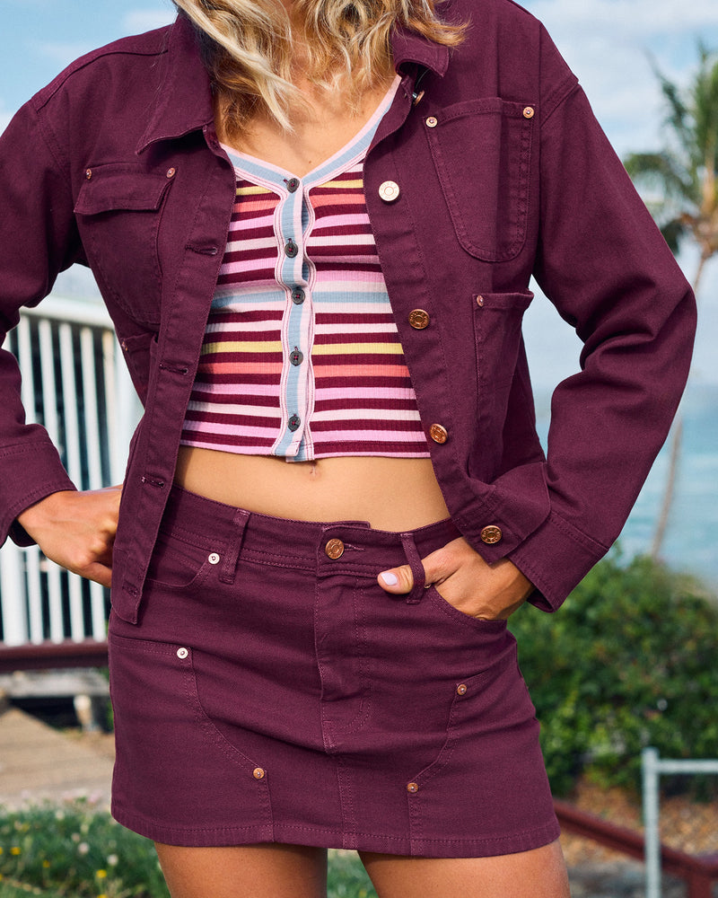 A person wearing a maroon denim jacket with carpenter details and a colorful striped crop top, paired with the Reel Life Skirt by Roxy, stands outdoors near a metal railing and greenery.
