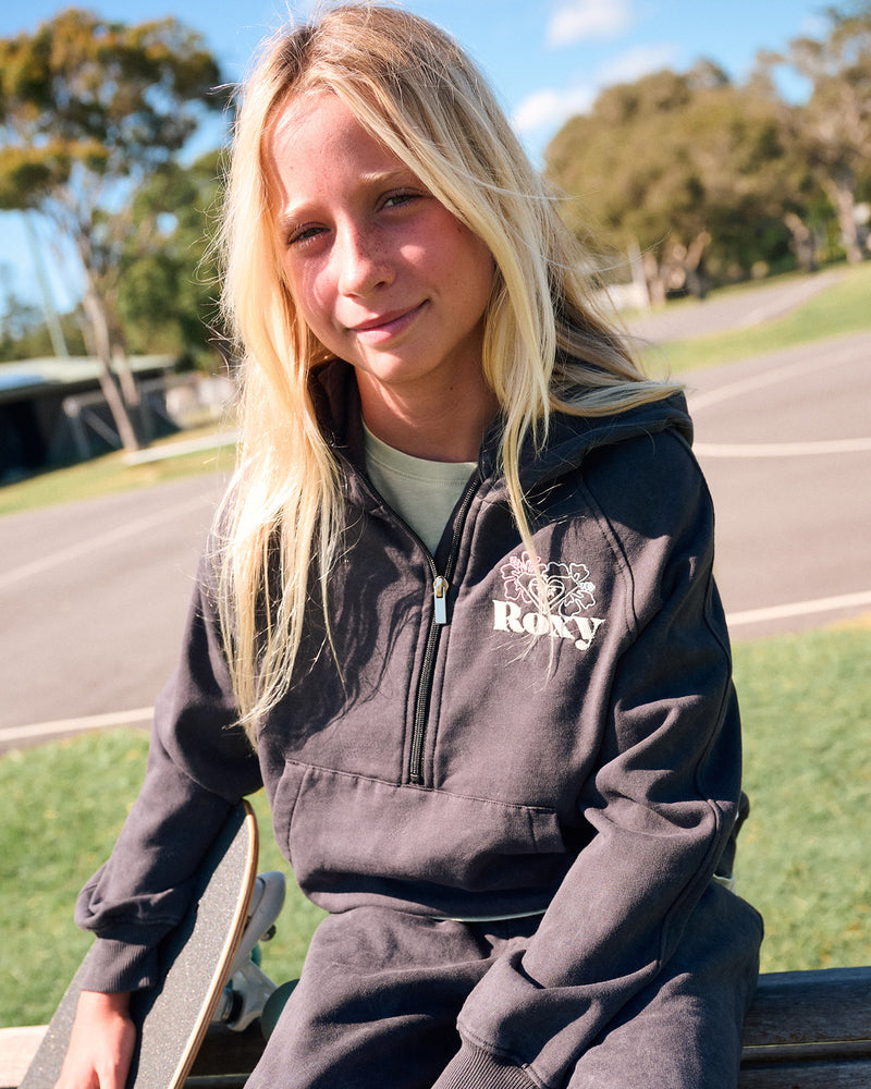 A smiling girl with long blonde hair wears a Roxy Girls' Heartbeat Half-Zip Hoodie in Phantom, featuring a kangaroo pocket, while holding a skateboard and sitting on a bench at a sunny outdoor skate park surrounded by trees.