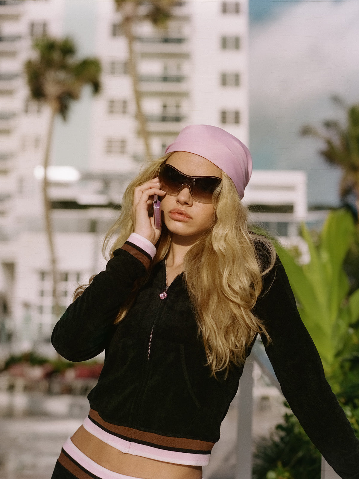 A woman with long blonde hair in a pink headscarf, large sunglasses, and the Roxy x Juicy Fitted Hoodie by Roxy channels Y2K style on her cellphone outdoors, set against a backdrop of a building and palm trees.