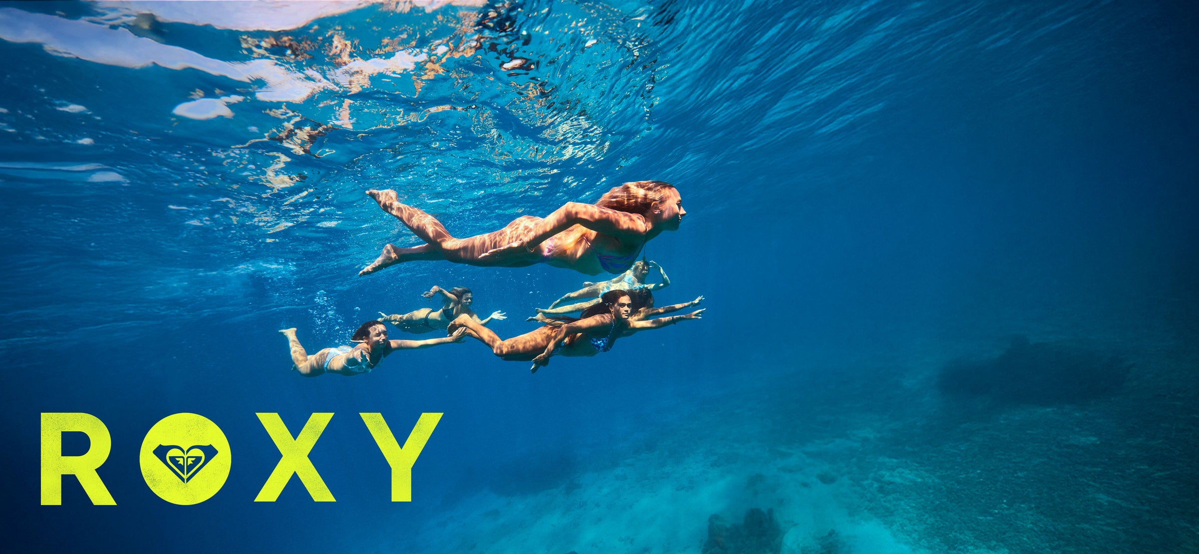 Three people swimming underwater with the Roxy logo above.