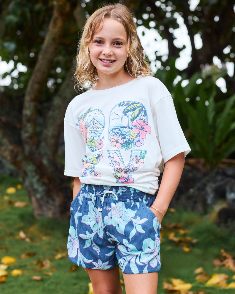 A smiling child with curly blond hair stands outdoors in a white t-shirt and Roxy Girls' Hula Soleil Short in Vintage Indigo, with green foliage and trees in the background.
