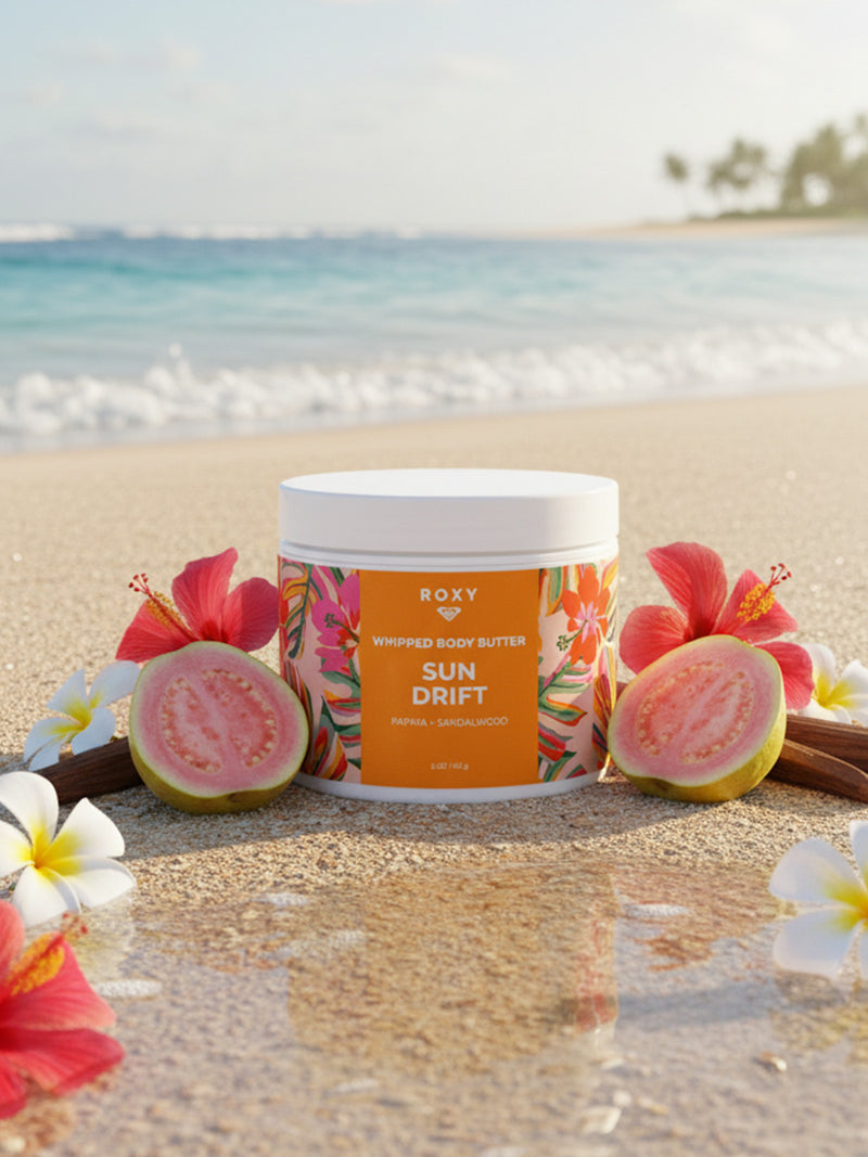 A jar of Roxy Sun Drift Body Butter rests on sandy beach surrounded by tropical flowers, guava, vanilla, and sandalwood—ideal for daily hydration and dry skin relief—with the ocean and palm trees softly blurred behind.