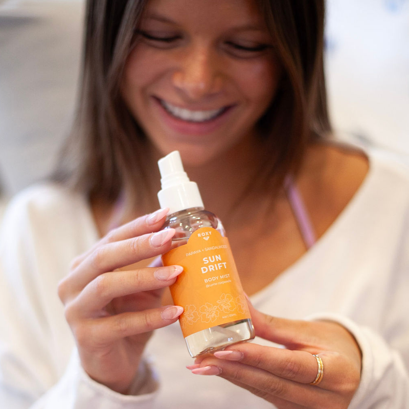 A smiling woman in a white top holds and looks at Roxy Sun Drift Body Mist, which features notes of hibiscus and comes in a spray bottle with an orange label adorned with a white floral design.