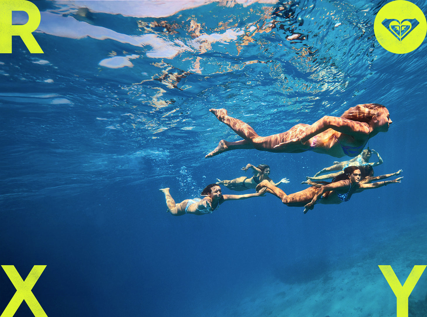 Three people swimming underwater with a clear blue ocean and Roxy logo.