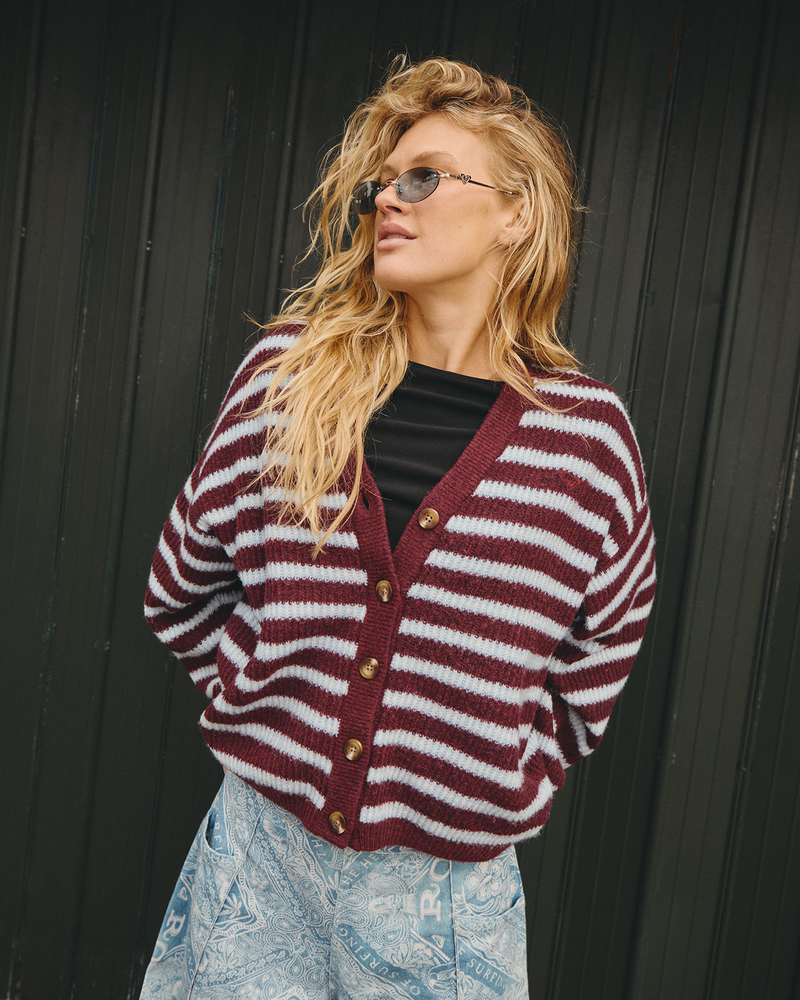A person wearing sunglasses, a relaxed Roxy Tamarindo Cardigan in maroon and white stripes layered over a black shirt, paired with a light blue patterned skirt, stands in front of a dark vertical panel wall and looks to the side—effortlessly combining style with lightweight warmth.
