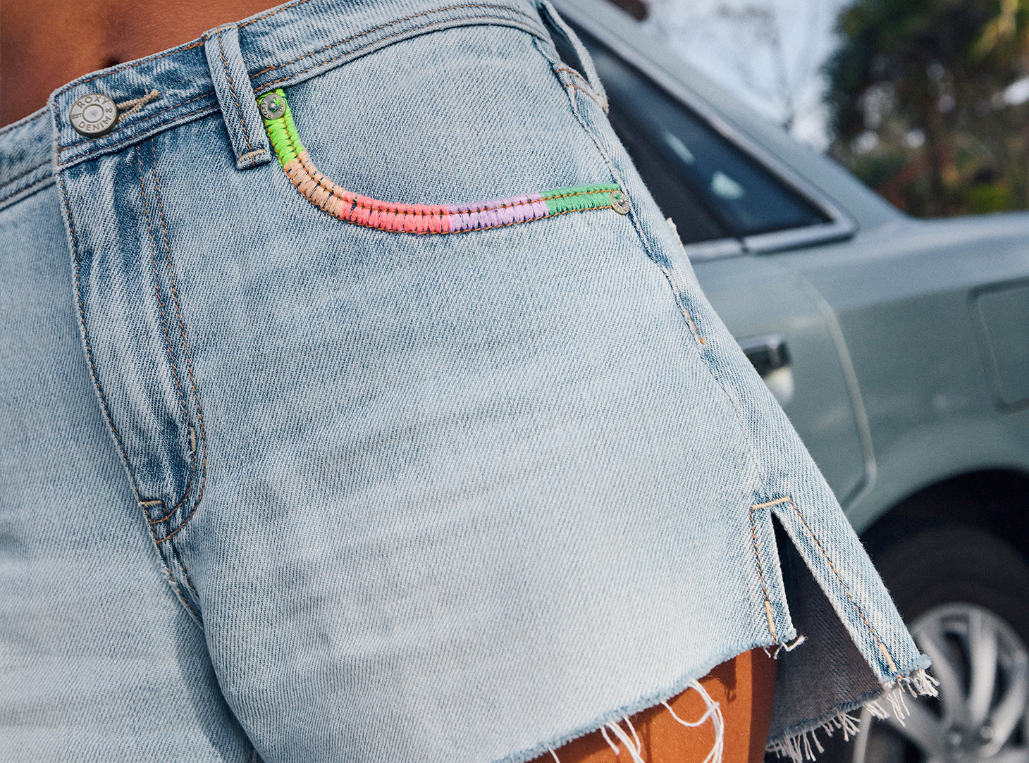 Close-up of a person wearing light-wash denim shorts with a colorful embroidered pocket detail, with a blurred car in the background.